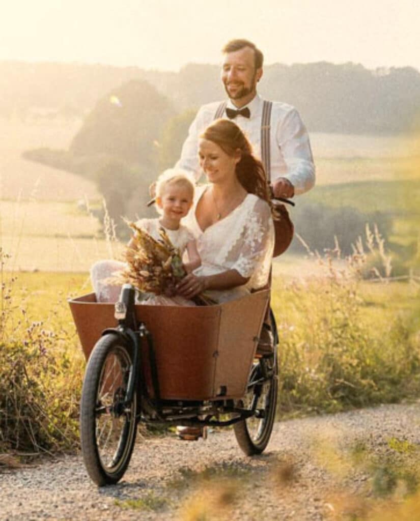 Brudeparret kører på en ladcykel med deres barn, der ser glad ud. En skøn måde at fejre kærligheden på.